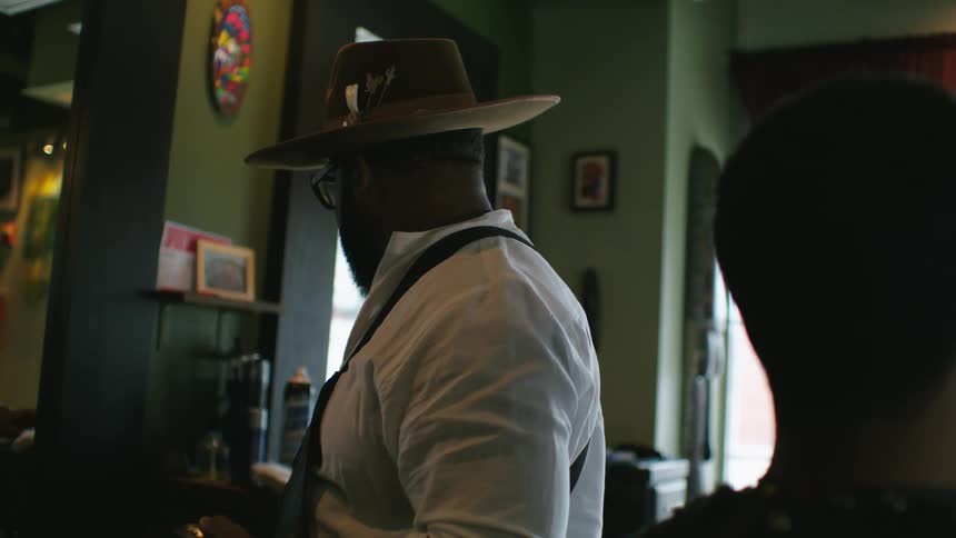 Barber with hat glasses beard white shirt and black apron trims mans hair with clipper in barber shop