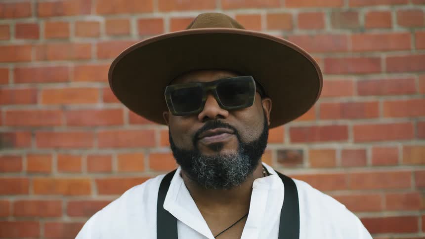 Bearded man wearing a hat and sunglasses looks up to the camera in front of a red brick wall