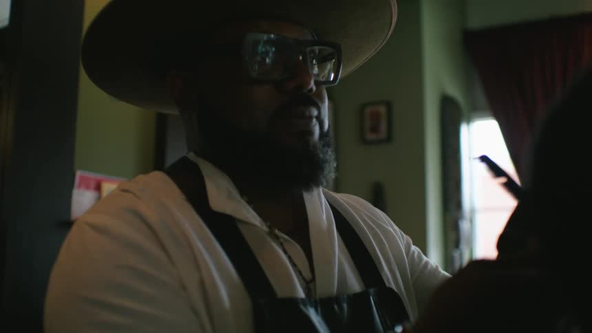 Barber with hat glasses beard white shirt and black apron cuts mans hair with clipper in barber shop