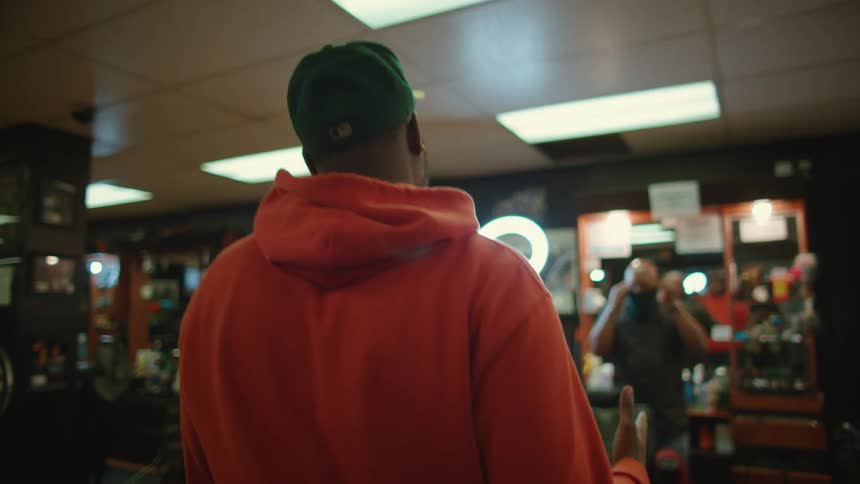 Man with orange hoodie green hat enters barber shop and shakes hands wit barber