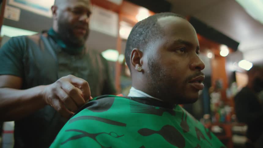 Barber throws cover over customer sitting in barber chair as client talks