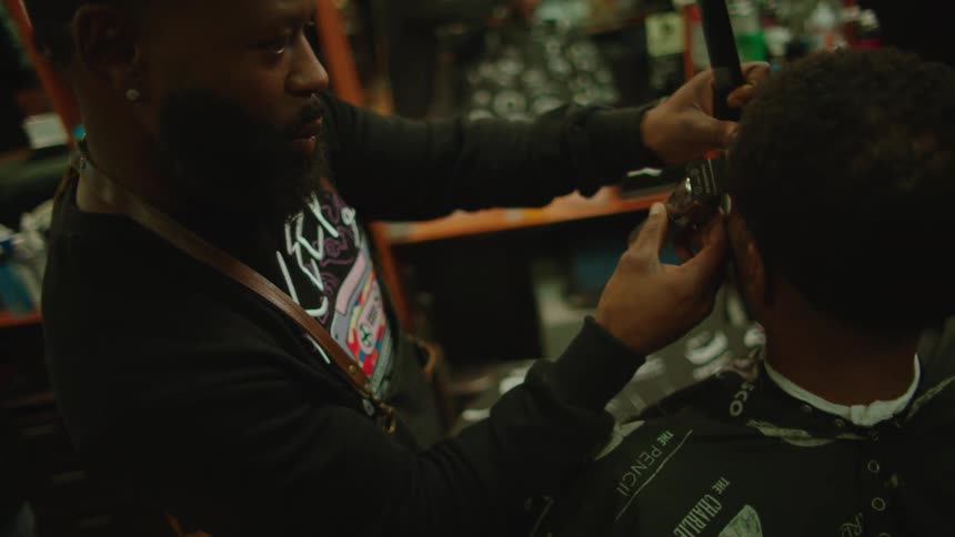Barber trims mans hair with hair clippers as another barber glances over