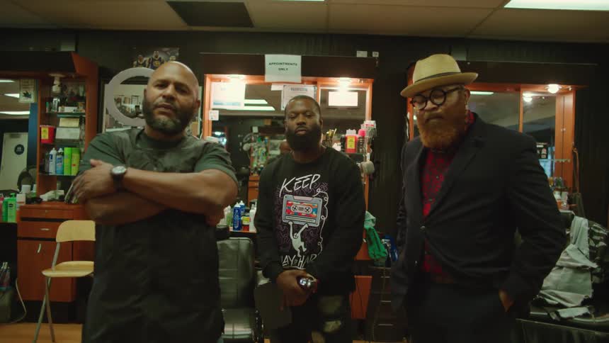 Three barbers with long beards apron shirt and jacket stand and look at camera in barbershop