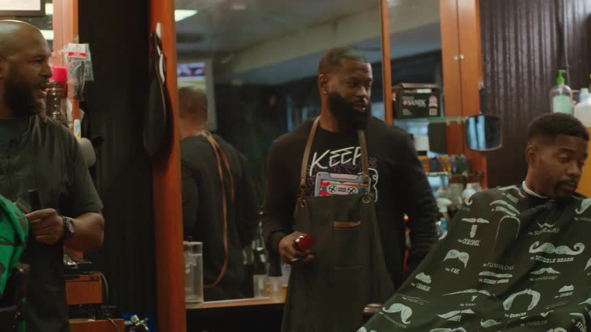 Barber with beard black sweatshirt and apron shows bald barber with beard smartphone and goes on to cut mans hair with hair clippers in barber shop