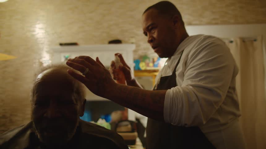An elderly man sits in a chair as a barber walks around him as he sprays his hair