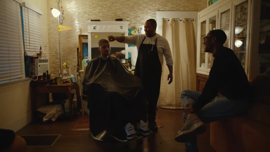 A man sits and laughs as a barber spins a man around in a chair and wipes his face off