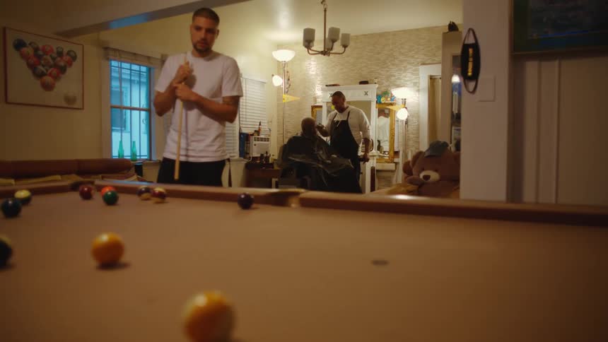 A man watches his opponent hit the cue ball on a pool table as a barber cuts a customers hair behind him