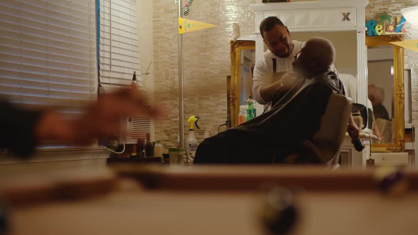 Man plays pool while another man has beard trimmed by barber while sitting in barber chair