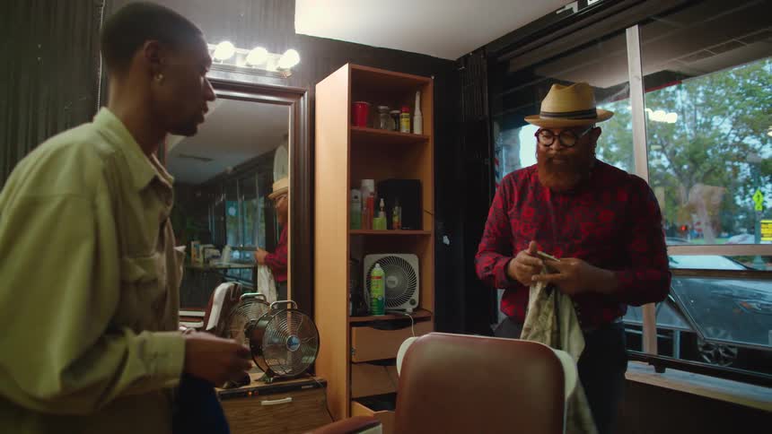 Man with earring and beige jacket pays barber with hat glasses beard and red shirt shaking hands in barber shop