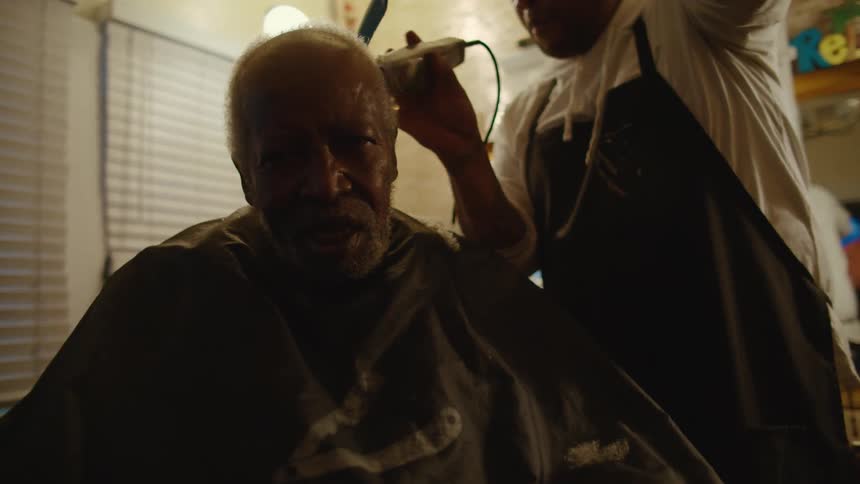 A male barber uses clippers to cut another mans hair in a small room