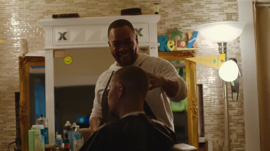 A male barber brushes a mans hair with a large round brush while another man watches them