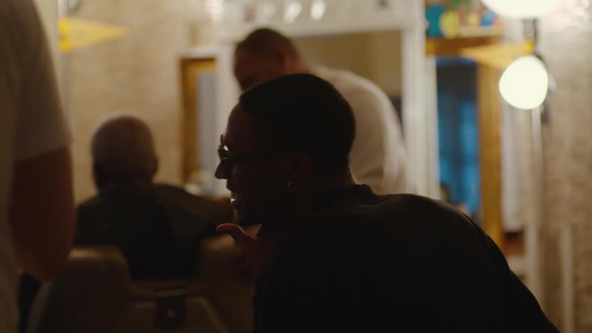 Two men laugh over bottle as barber looks over from his customer and also laughs