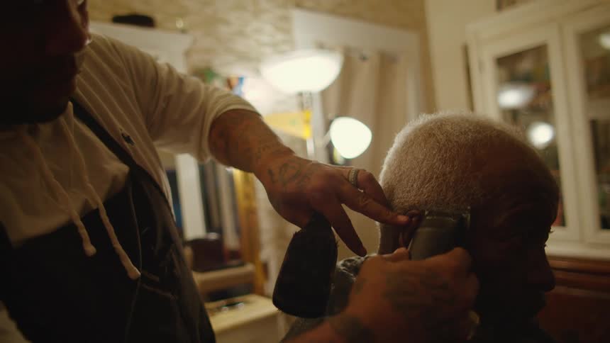 Barber with white sweatshirt black apron cuts mans hair with clipper in barber shop