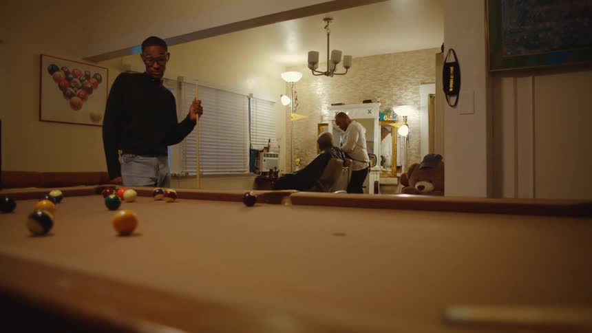 Two men with glasses and dark sweater play pool while barber cuts customers hair in the background