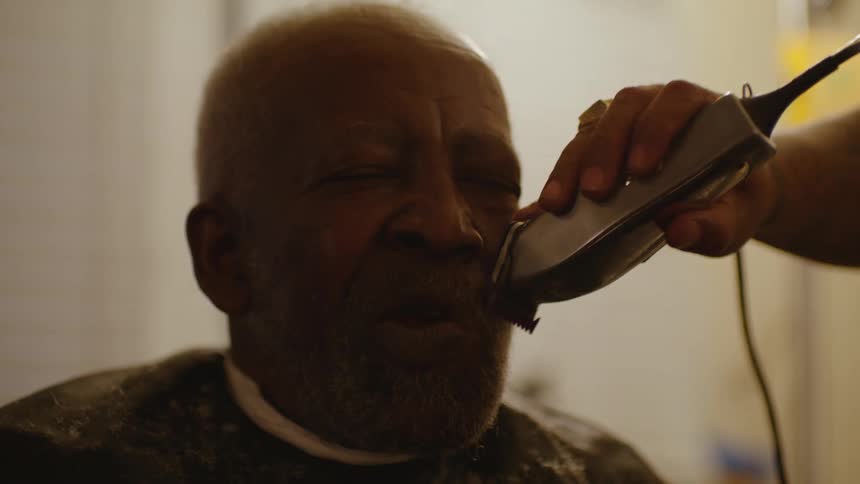 Barber with white shirt and black apron shaves middle aged man with clipper in barber shop