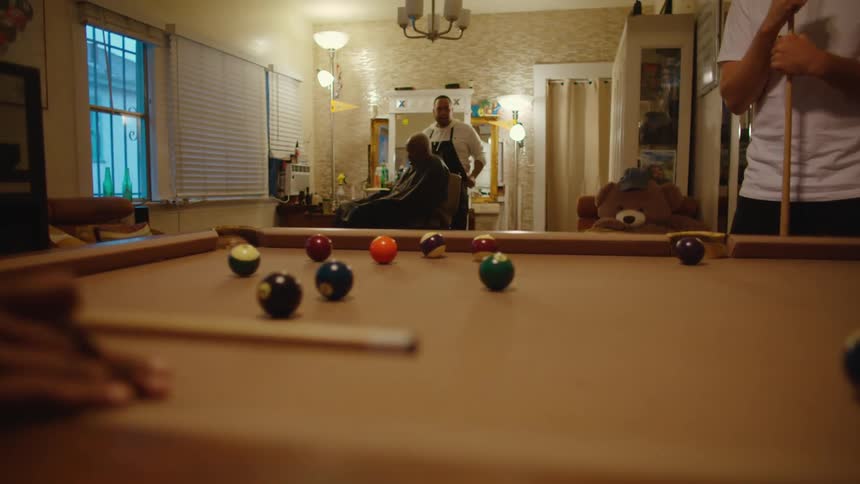 Barber with white shirt and black apron shaves middle aged man with clippers while a group of men plays pool in barbershop