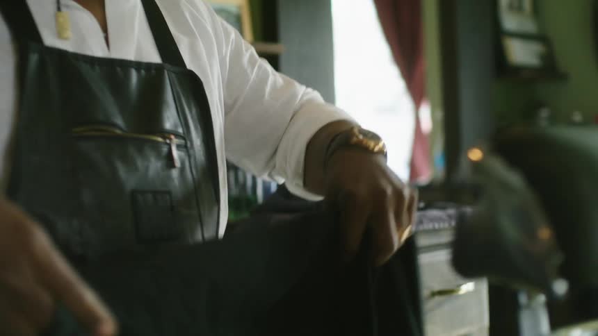 Barber with brown hat glasses and beard with white shirt and black apron folds apron and puts in chair in barber shop