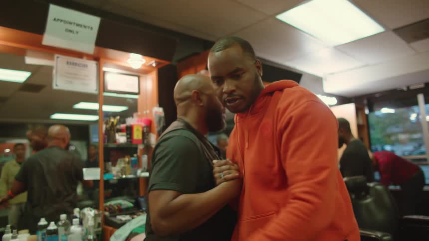 A customer in a barber shop raises from the barber chair to greet the barber and another customer