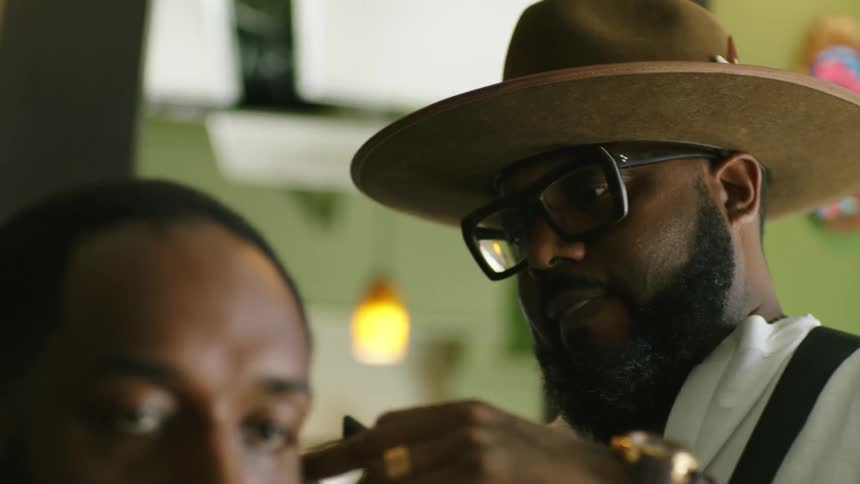 A man wearing a hat is cutting the hair of a man sitting down