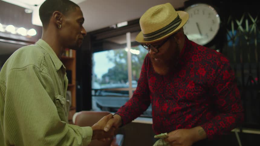 Man with earrings and beige jacket pays barber with hat and red shirt in barber shop