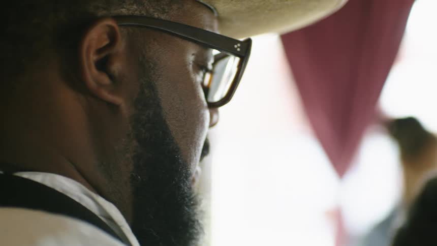 Barber with hat glasses and beard cuts customers hair in barber shop