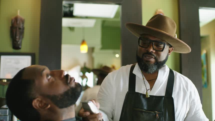 Barber with brown cowboy hat glasses beard white sweatshirt and black apron shaves mans beard with clippers in barber shop