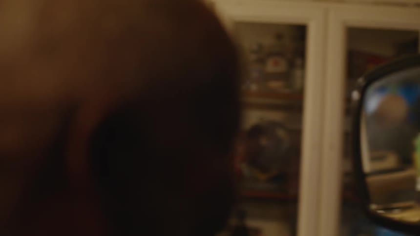 An elderly man sitting in a barbers chair smiles at his own reflection in a handheld mirror