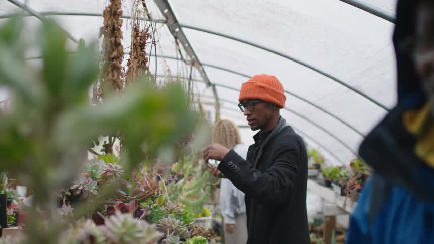 Man wearing jacket in greenhouse talks while man in background takes down dried cuttings