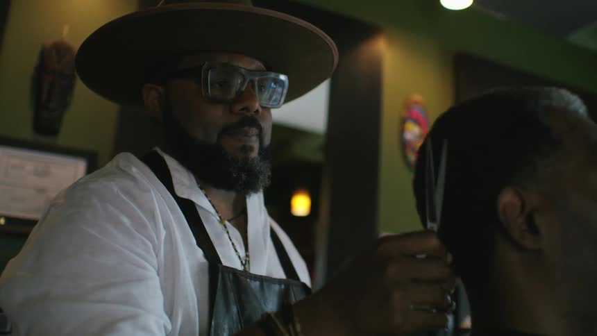 Barber with hat glasses beard white shirt and apron cuts mans hair with scissors in barber shop