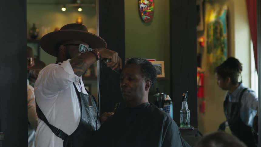Barber wearing a hat shaves the sideburns of a client sitting on a chair in the barber shop