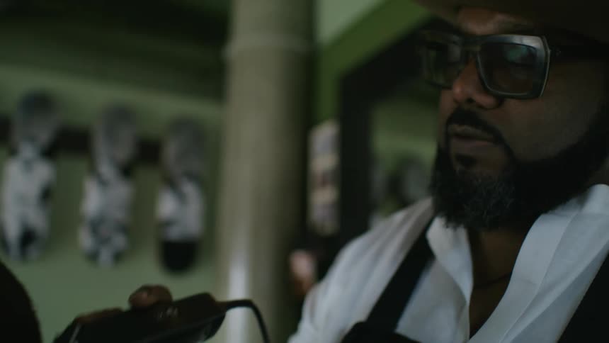 Barber with brown hat glasses beard white shirt and black apron cuts mans hair with clipper in barber shop