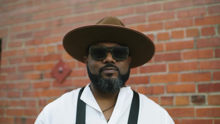 Bearded man wearing a hat and sunglasses stands with an apron in front of a red brick wall in the city