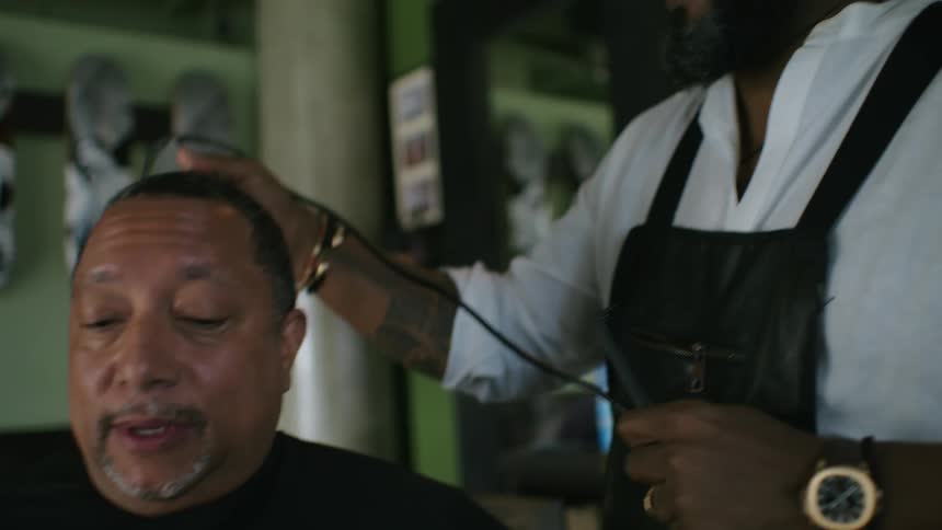Barber with brown cowboy hat glasses beard white sweatshirt and black apron cuts mans hair with clipper in barber shop