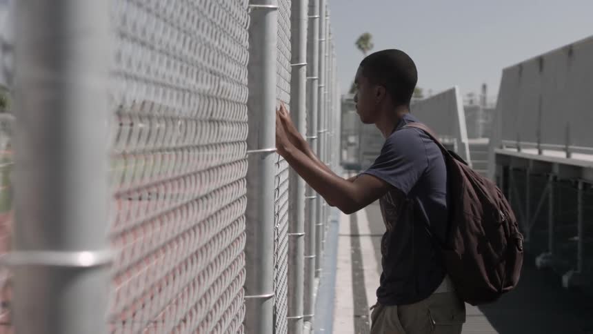 Young man wearing a backpack standing while holding and looking through a fence