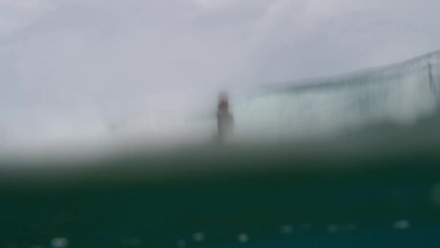 A surfer wearing shorts and a dark colored shirt rides a wave and the view changes to underwater below