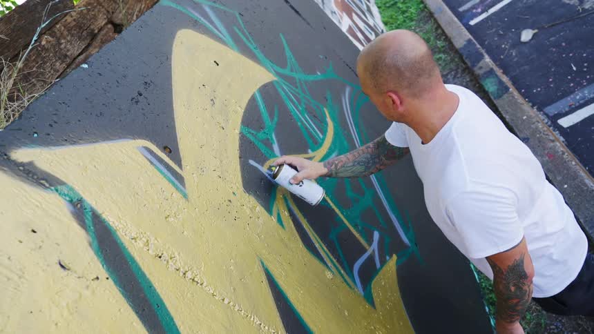 A tattooed man spray painting graffiti onto a concrete wall