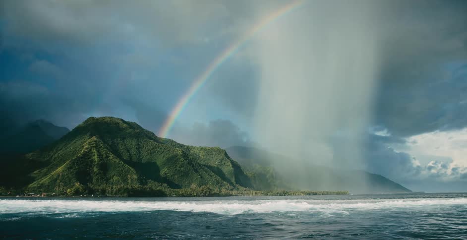 Rainbow curves over vegetation covered mountain and crashing waves near shore