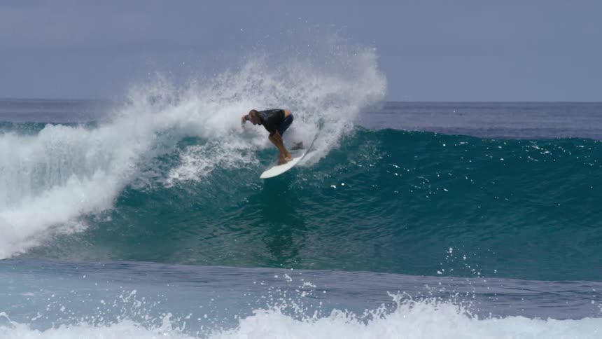 A surfer wearing shorts and a dark colored shirt rides a big wave as water splashes behind him
