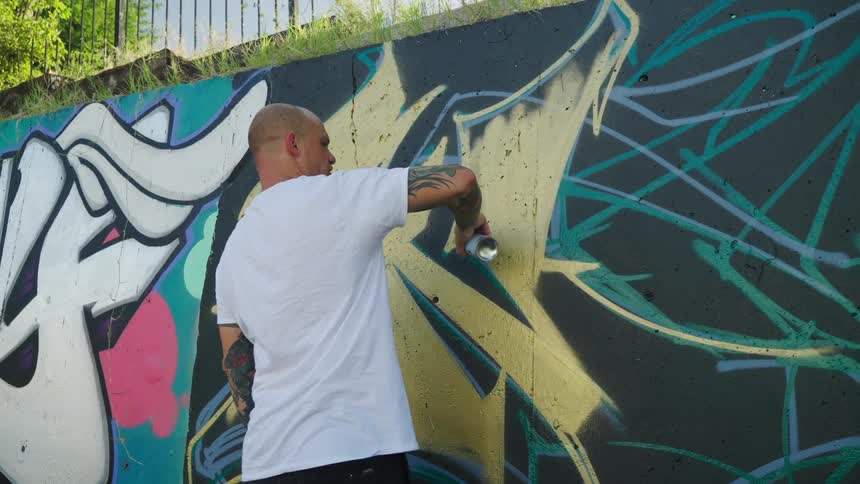 Man with tattooed arms spraying graffiti on a wall in the city at morning