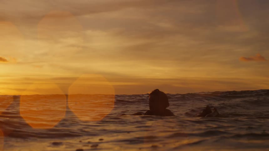 A surfer paddles through gentle waves under an orange sky