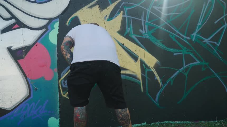 Tattooed artist spraying graffiti on the wall on a town at morning