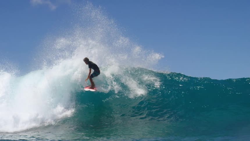 A surfer wearing shorts and a tshirt rides a big wave