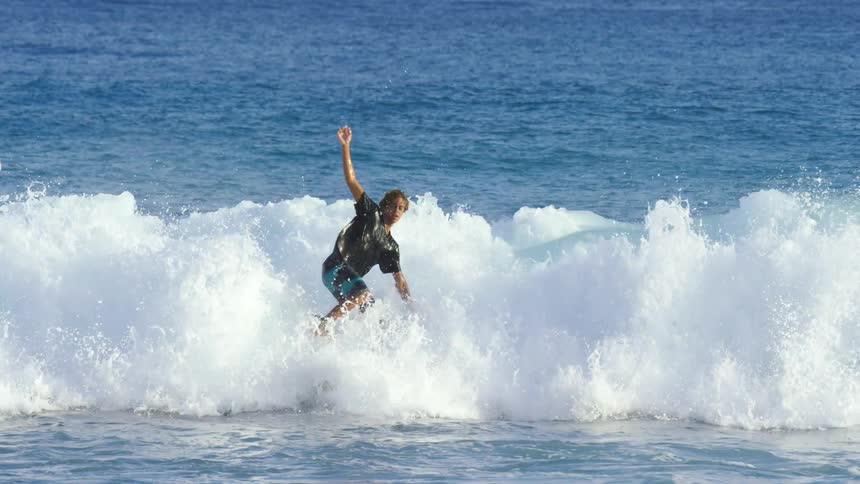 A surfer wearing shorts and a tshirt falls into the water as he rides a wave