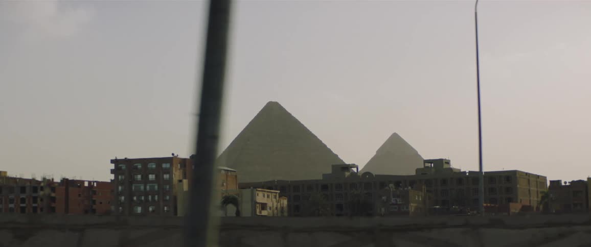 Van drives over bridge and along road passing apartment buildings backed by pyramids