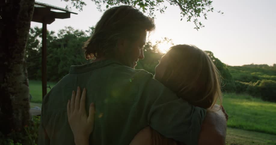 A man and woman embrace and kiss while watching the sun descend behind a woodland