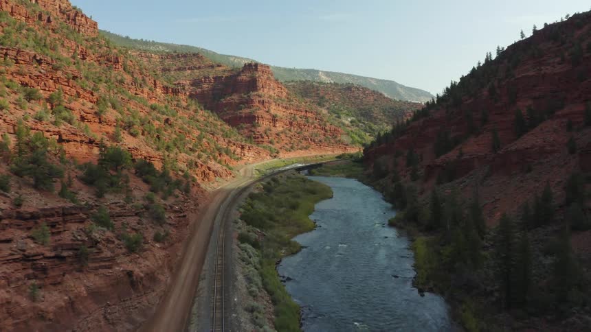 A river runs alongside a railway and dirt road along the floor of a narrow mountain valley during the day