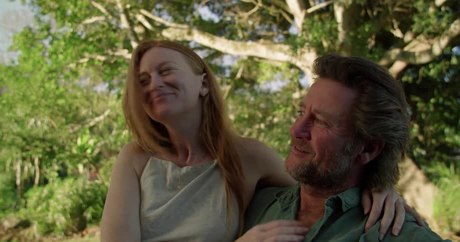 Man with beard and blue shirt holds woman with red hair and grey vest sitting in garden lovingly looking into each others eyes and making out
