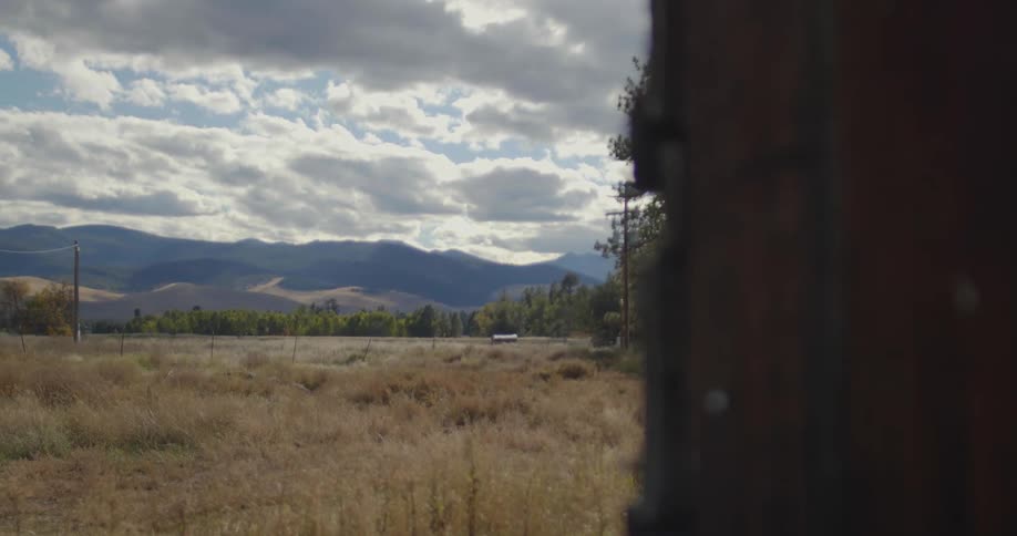Farm house sits in middle of plot bordering water near mountains under a cloudy sky