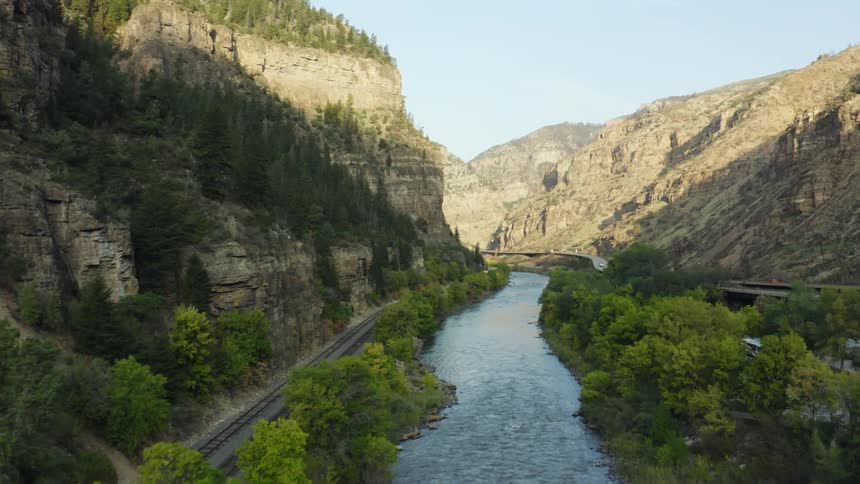 A river running alongside a road and a railway on the floor of a canyon on a sunny day