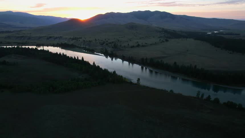 A river running through a hilly countryside as the sun begins to peak over mountains in the distance